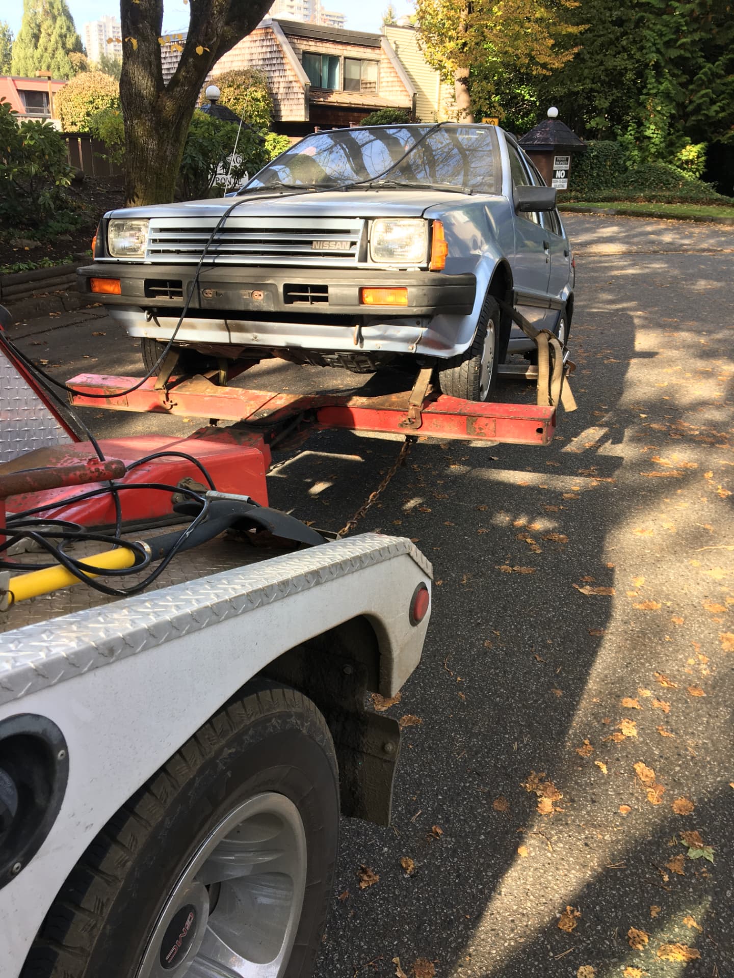 An older blue Nissan hatchback strapped down on the tow deck on a residential street